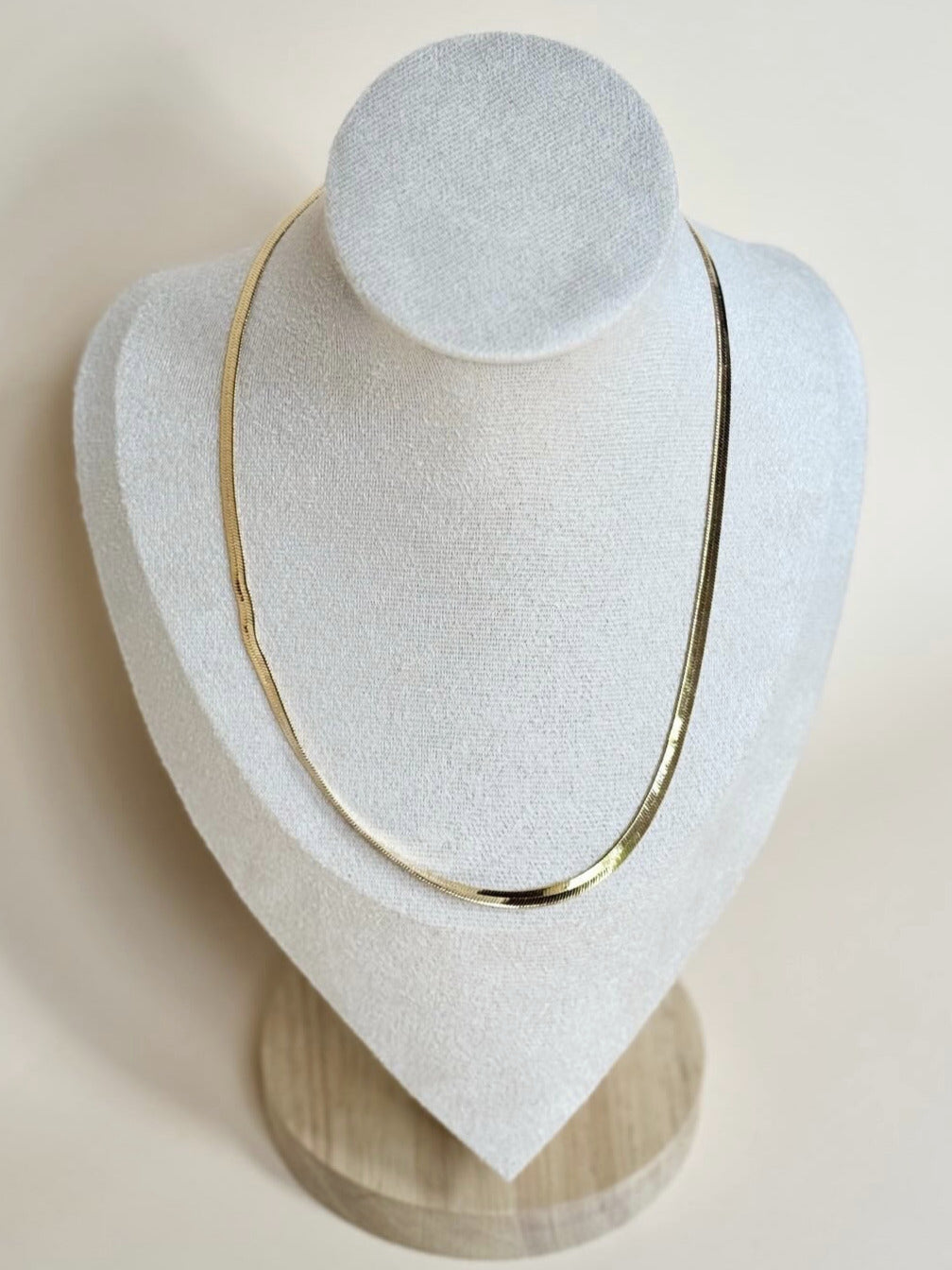 A gold-filled herringbone necklace displayed on a white stand with a wooden base.