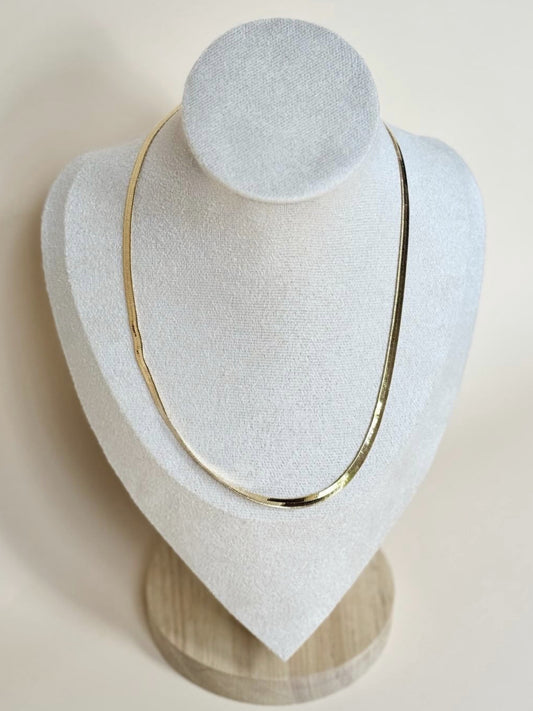A gold-filled herringbone necklace displayed on a white stand with a wooden base.
