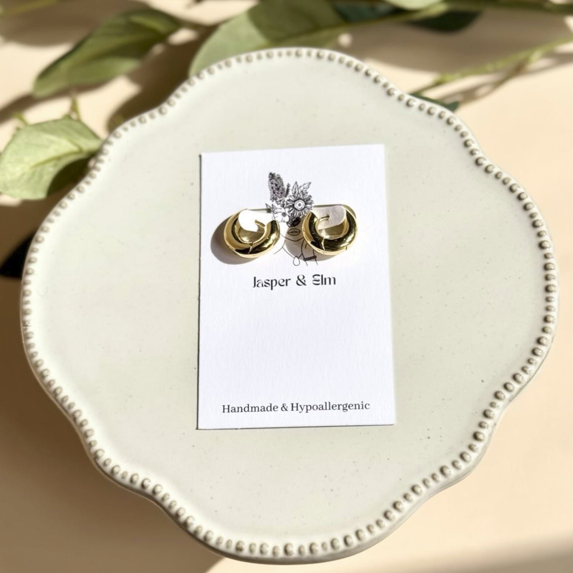 A pair of large 18k PVD gold-plated hoop earrings displayed on a white plate with a brand card behind them.