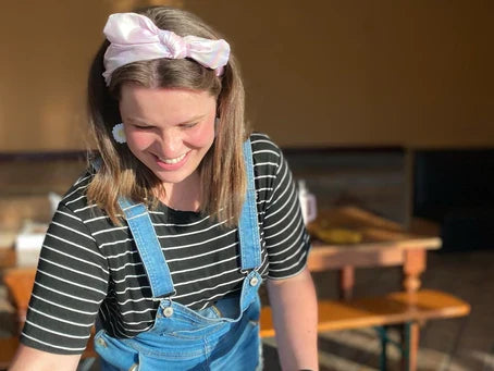 A cheerful woman wearing denim overalls, a striped shirt, and a pink headband smiles brightly in a sunlit, casual setting.