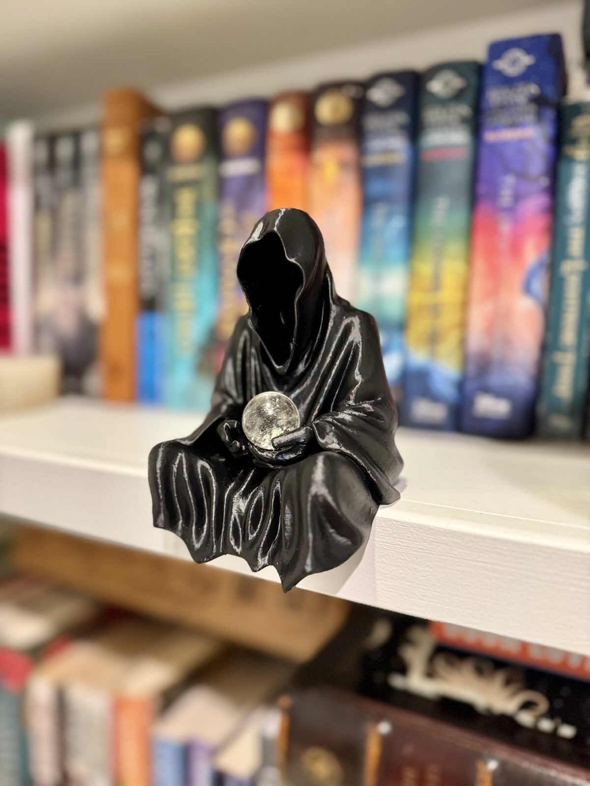 Statue of a hooded figure holding a crystal ball on a bookshelf with books in the background